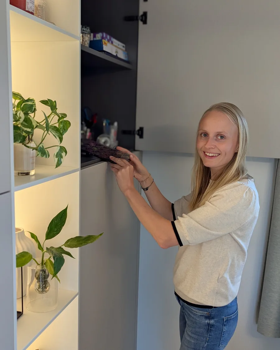 Coralie Poncelet sourit en rangeant le haut d'un placard gris, à côté de niches éclairées garnies de plantes