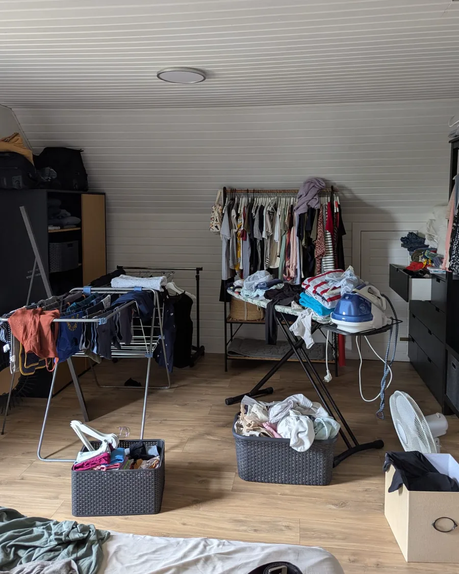 Dressing sous combles avant rangement : étendoirs chargés, planche à repasser, paniers et cartons au milieu du sol